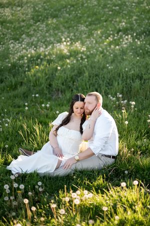 Couple sitting closely together in a green meadow filled with dandelions during a warm sunny engagement session.
