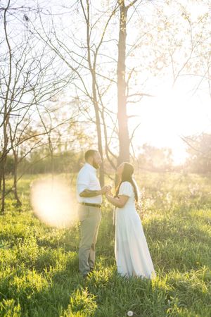 Couple holding hands in a sunlit forest clearing with golden backlight streaming through bare trees in spring.