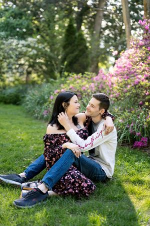 Couple sitting together on green grass in front of colorful flowering bushes in a beautiful garden.