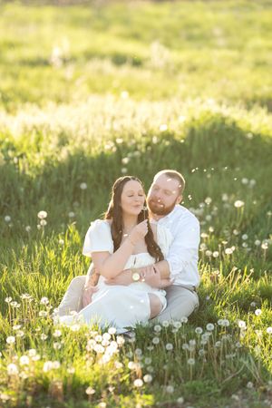 Couple sitting in sunlit dandelion field, woman in white dress blowing dandelion seeds in golden hour light.