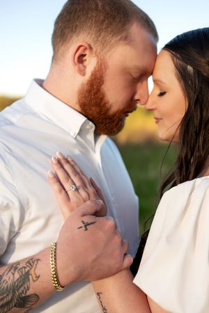 Close-up of couple touching foreheads tenderly, showing engagement ring and cross tattoo in warm outdoor light.