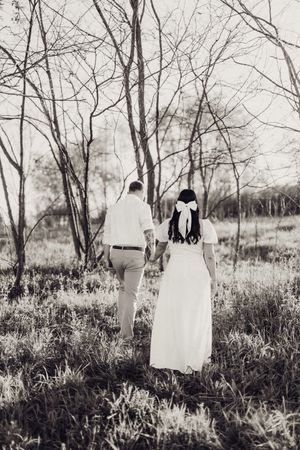 Black and white photo of a couple holding hands walking through a serene forest in wedding attire.