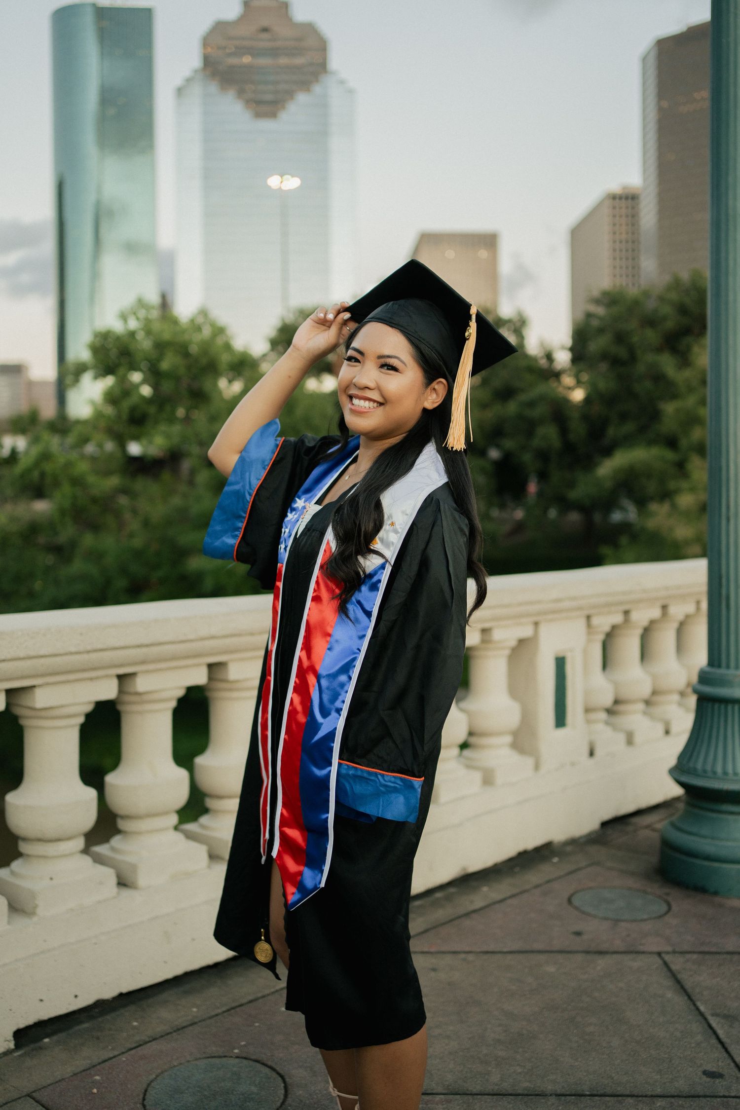 A graduate in cap and gown poses cheerfully against a cityscape backdrop.