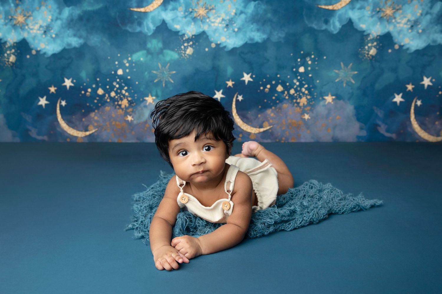 Baby in starry outfit poses on blue backdrop with celestial moon and stars design.