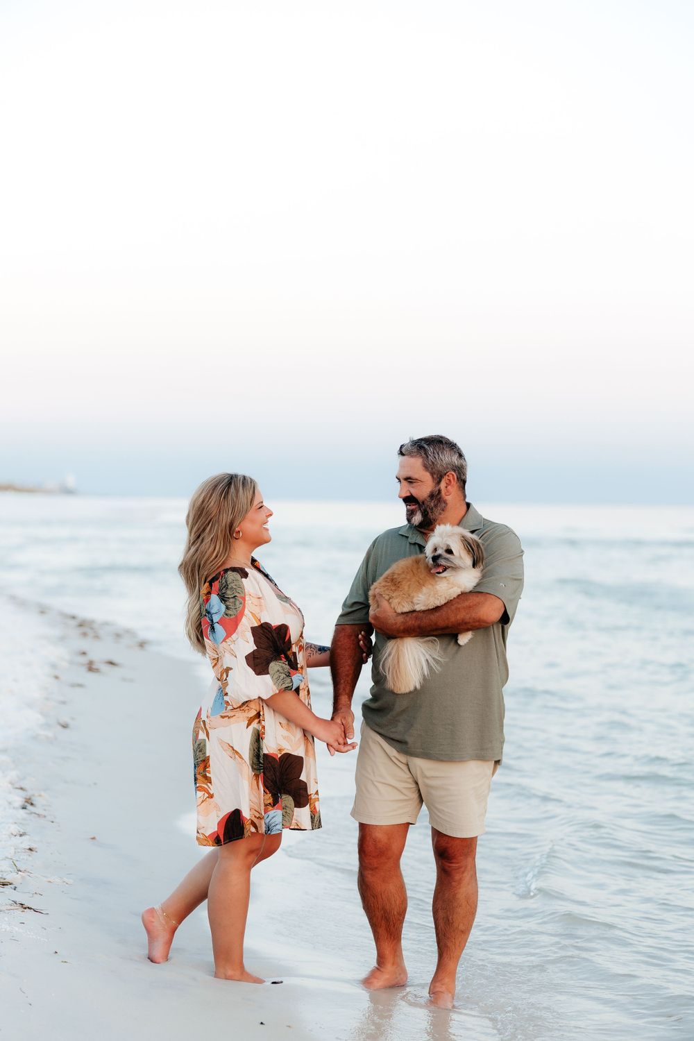 A couple stands on a beach at sunset holding their small dogs while enjoying the ocean view.