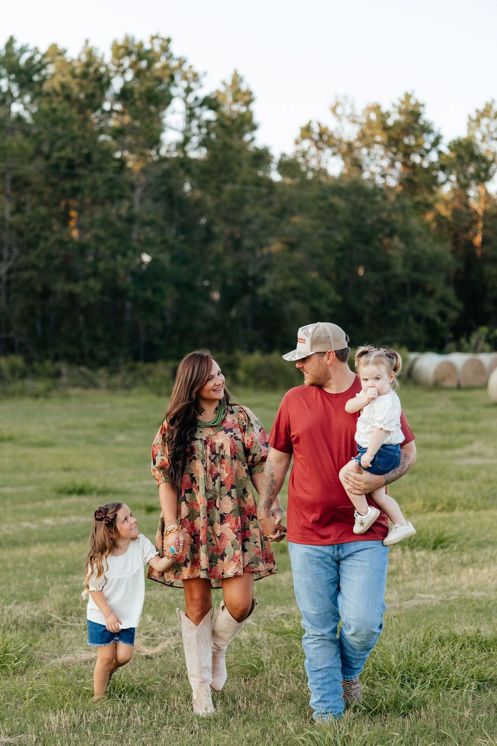 A family walks together in a grassy field with hay bales and trees in the background at sunset.
