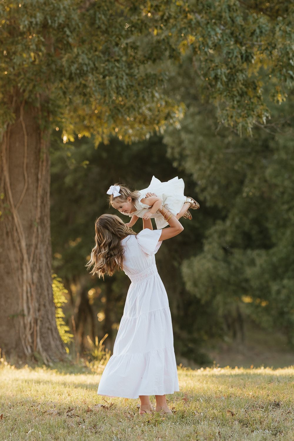 A mother in a white dress lifts her young daughter in the air outdoors near a large tree in golden sunlight.