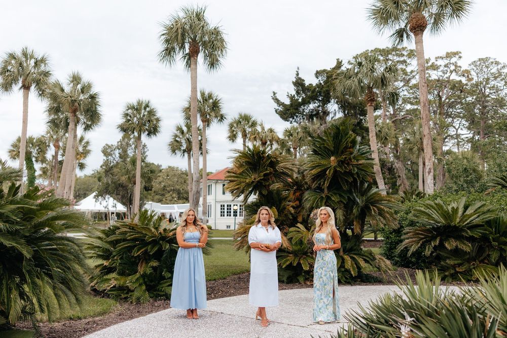 Three bridesmaids in blue and floral dresses stand on a garden path surrounded by palm trees at a Florida wedding venue.