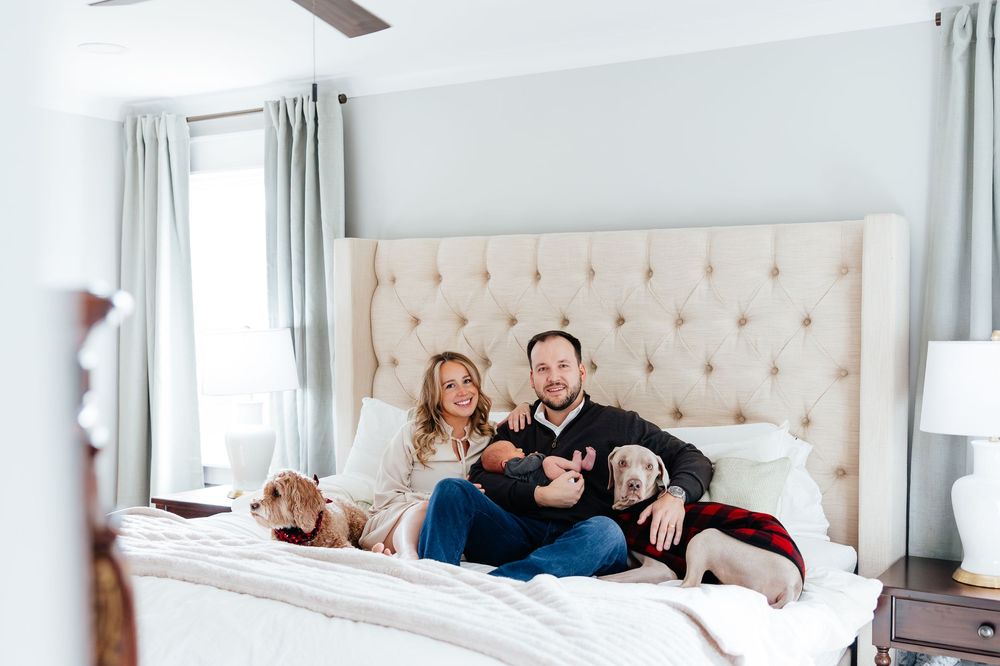 Couple with newborn baby and two dogs relaxing on a tufted beige bed in a bright, modern bedroom.