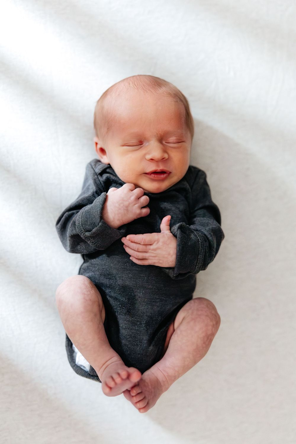 Newborn baby sleeping peacefully in a dark gray onesie, lying on a white blanket with hands folded on chest.