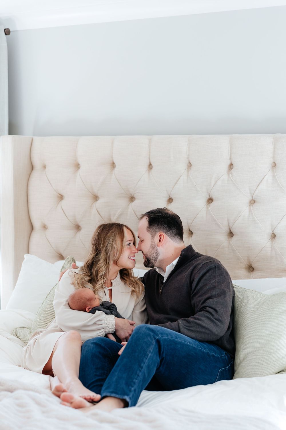 A couple sits on a bed gazing at each other while holding their newborn baby in a cozy bedroom.
