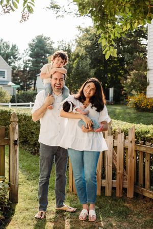A family laughs joyfully together by a wooden fence in their backyard garden on a sunny summer day.