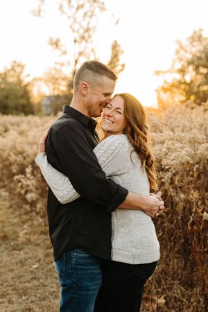 A loving couple embraces and laughs together during golden hour in a field of tall grass at sunset.