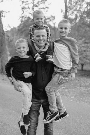 Black and white candid portrait of a parent playfully carrying three young children outdoors.