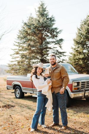 Couple stands together outdoors in casual fall attire next to a vintage pickup truck with evergreen trees in background.