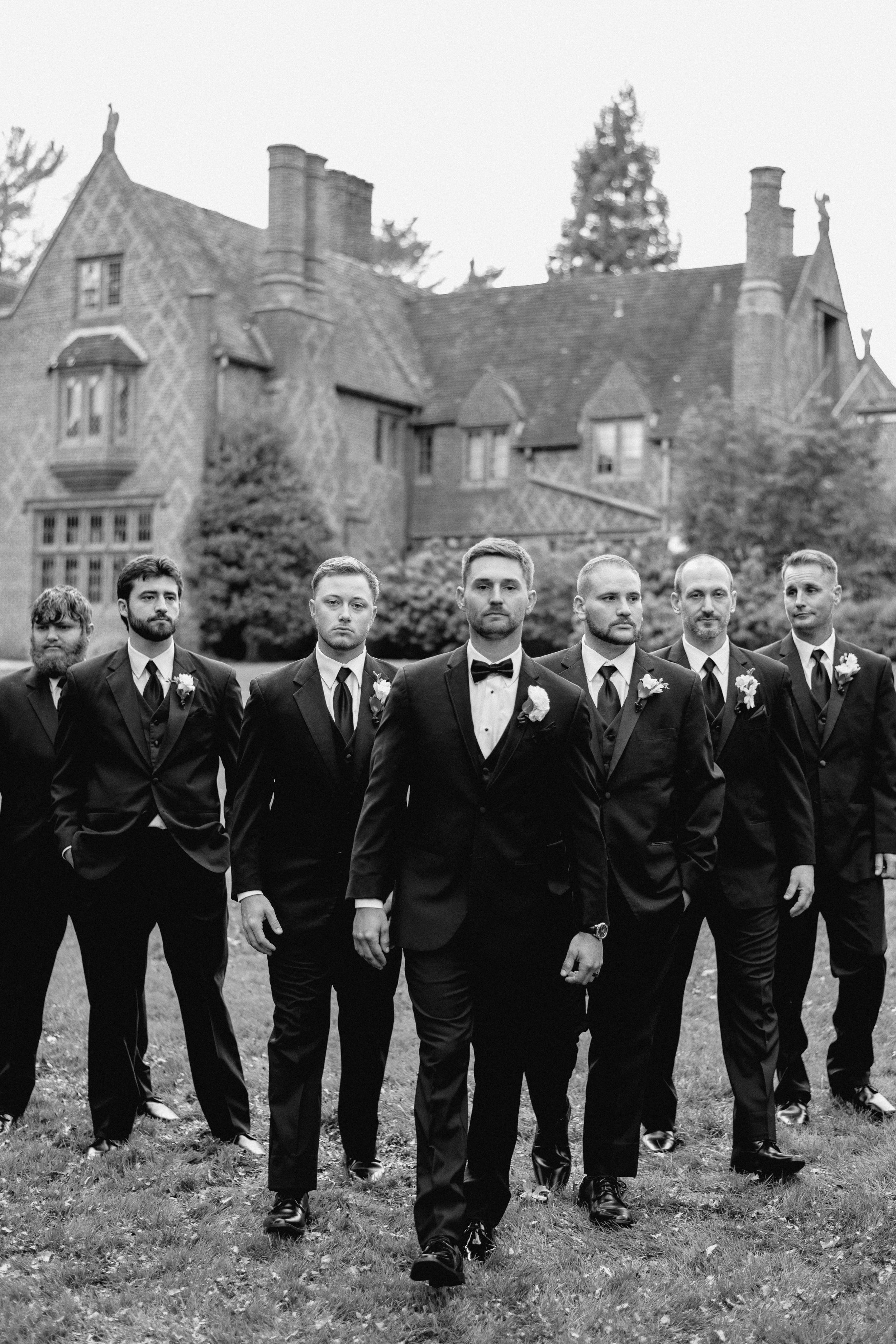 Groomsmen in dark suits pose in front of a Gothic-style mansion.
