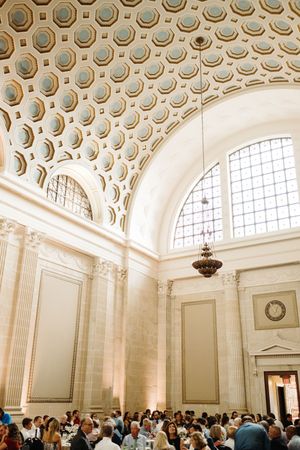 Ornate domed ceiling with circular coffers and arched windows in a grand hall filled with visitors beneath a chandelier.