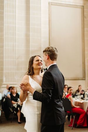 Newlyweds share their first dance together in an elegant ballroom with classical white pillars and reception tables.