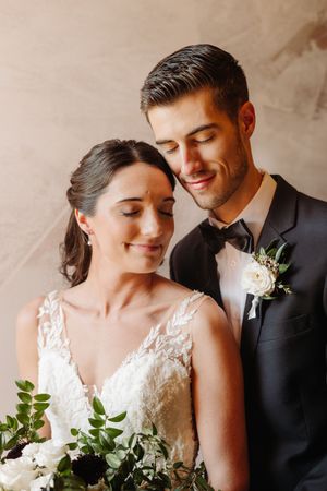 Romantic wedding portrait of newlyweds sharing an intimate moment with greenery bouquet and classic black tuxedo and lace gown.