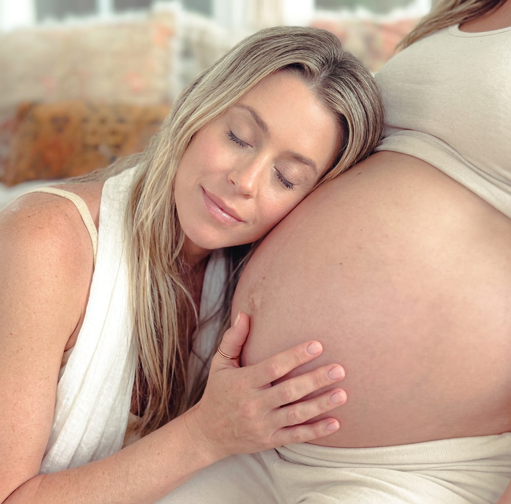 Pregnant woman resting her head on another person's baby bump, sharing a tender bonding moment.