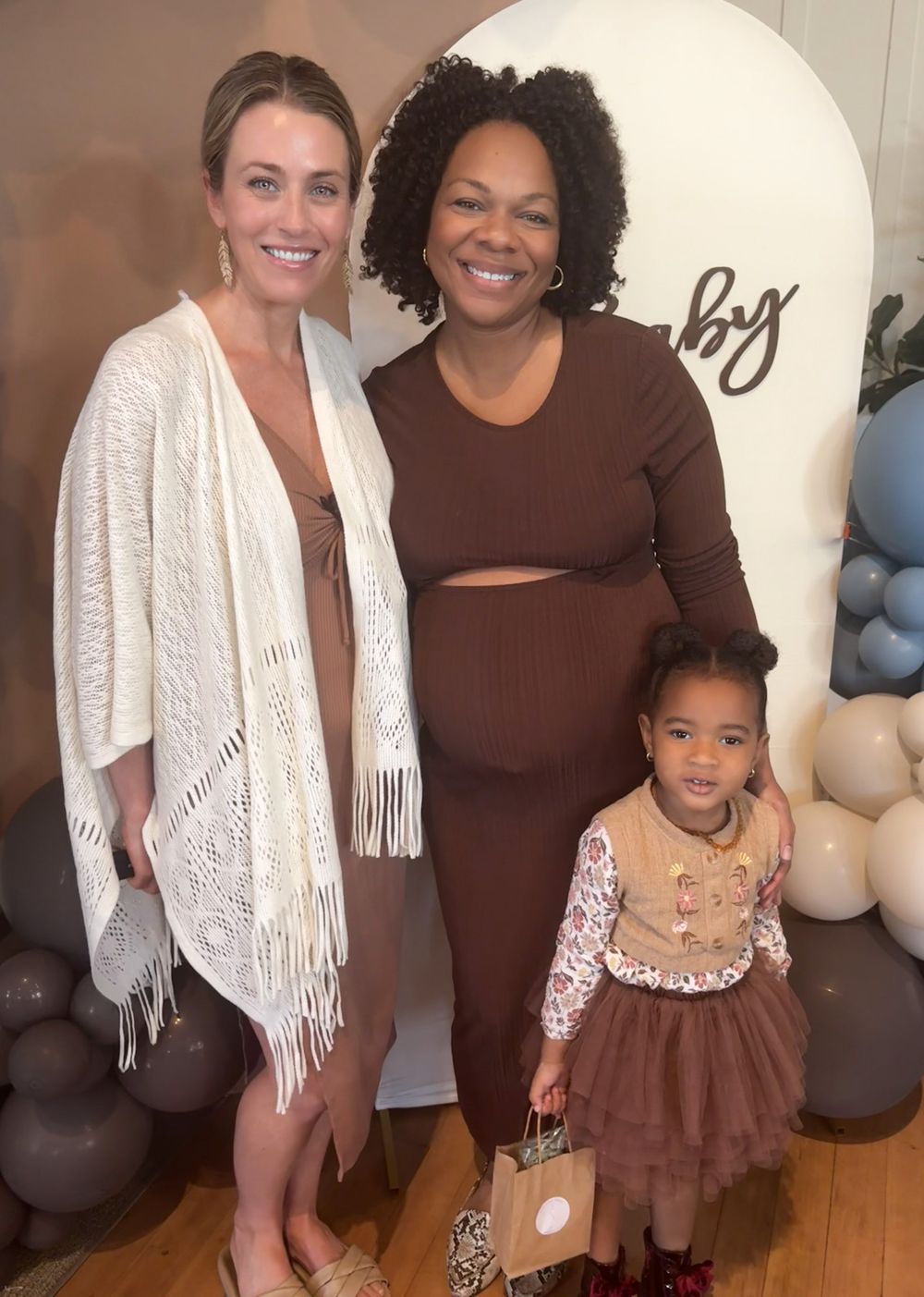 Two women and a toddler girl pose together at a baby shower with balloon decorations.