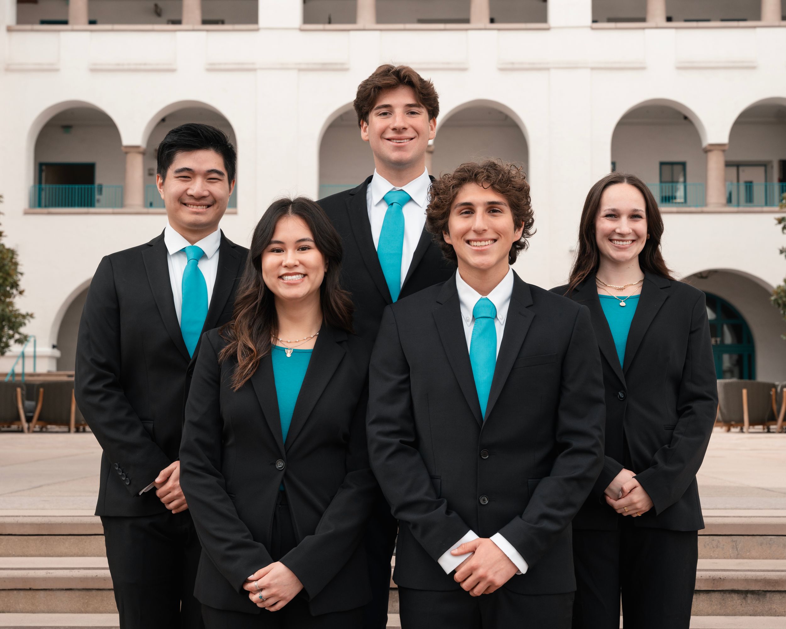 Group photo of graduates wearing black suits with turquoise ties standing in front of a white building.