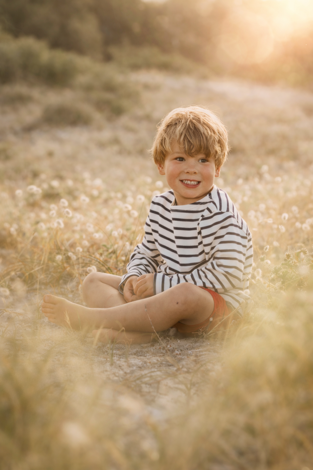 Child in striped shirt sits in sunlit field of wildflowers during golden hour.