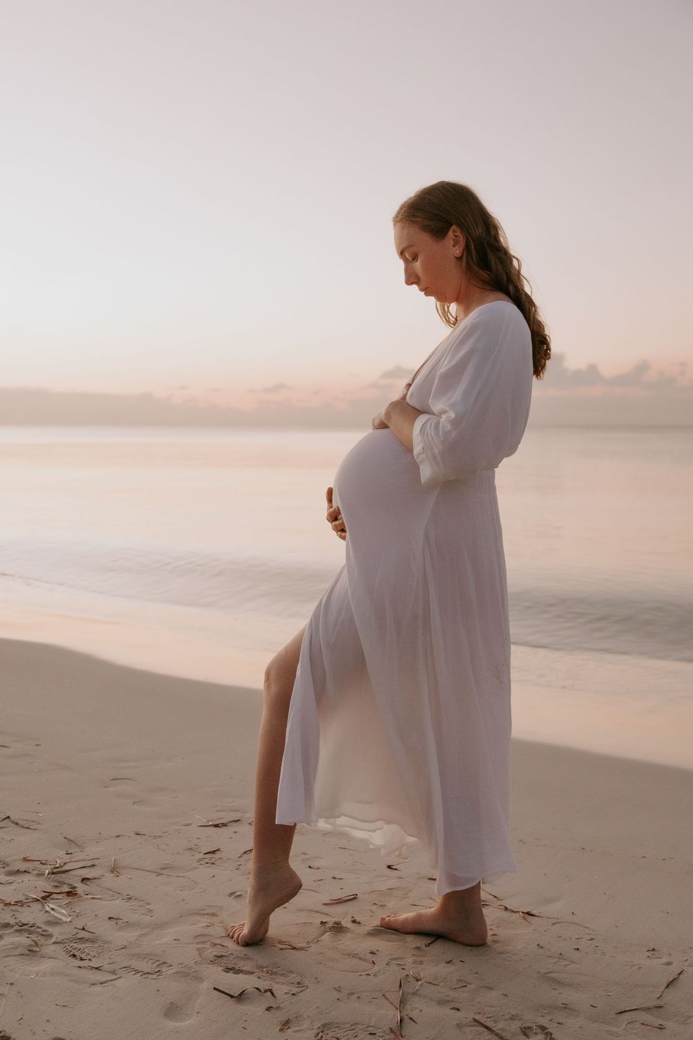 Pregnant woman in white dress holding her belly on a sandy beach at sunset.