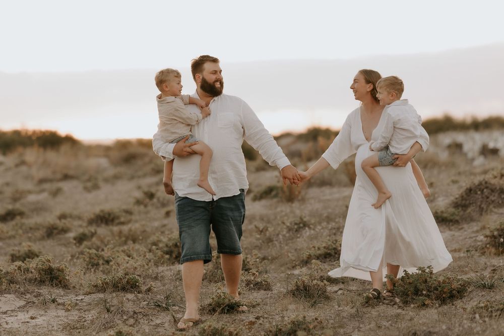 A family of four walks hand-in-hand on a sandy beach, parents each holding a young child at golden hour.