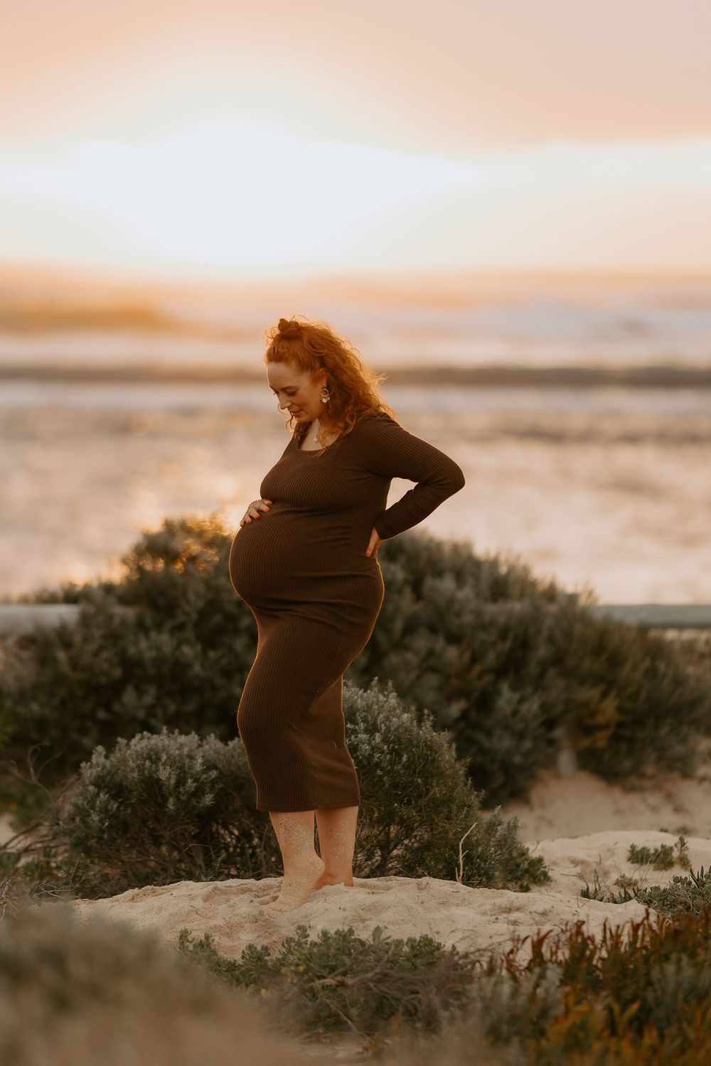 Pregnant woman in brown dress standing barefoot on sandy beach at sunset, cradling her baby bump with curly red hair.