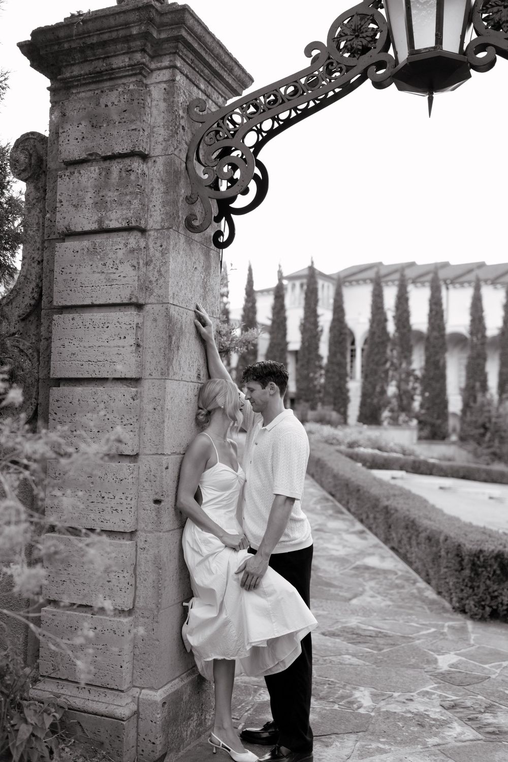 A romantic black and white photo of a couple embracing by a stone pillar with cypress trees in the background.