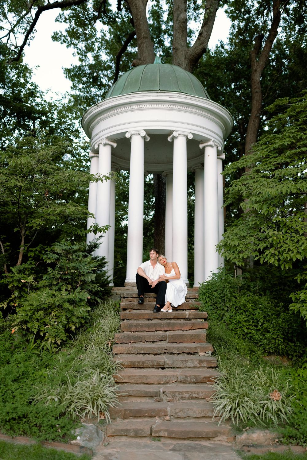 A white gazebo with classical columns sits atop stone steps surrounded by lush green trees and landscaping.