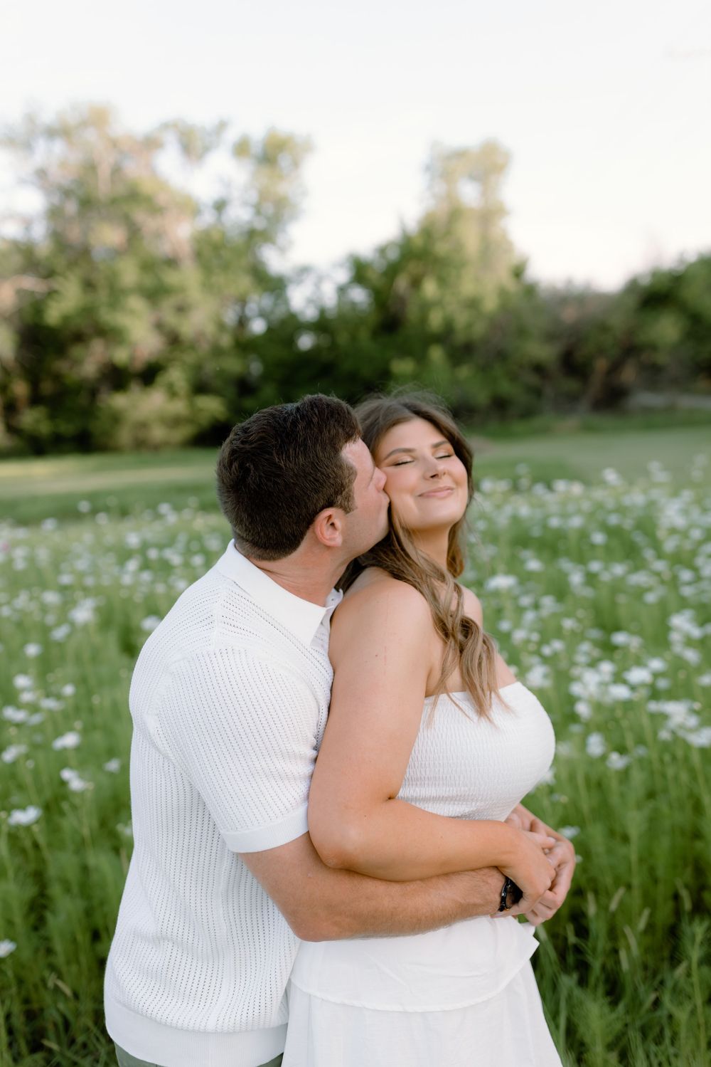 A couple shares tender moments in a blooming wildflower meadow wearing white outfits on a sunny day.