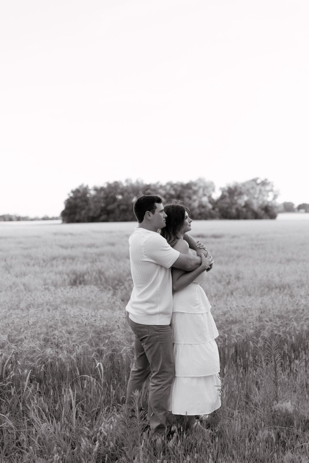 A couple embraces lovingly in a rustic field setting captured in black and white photography.