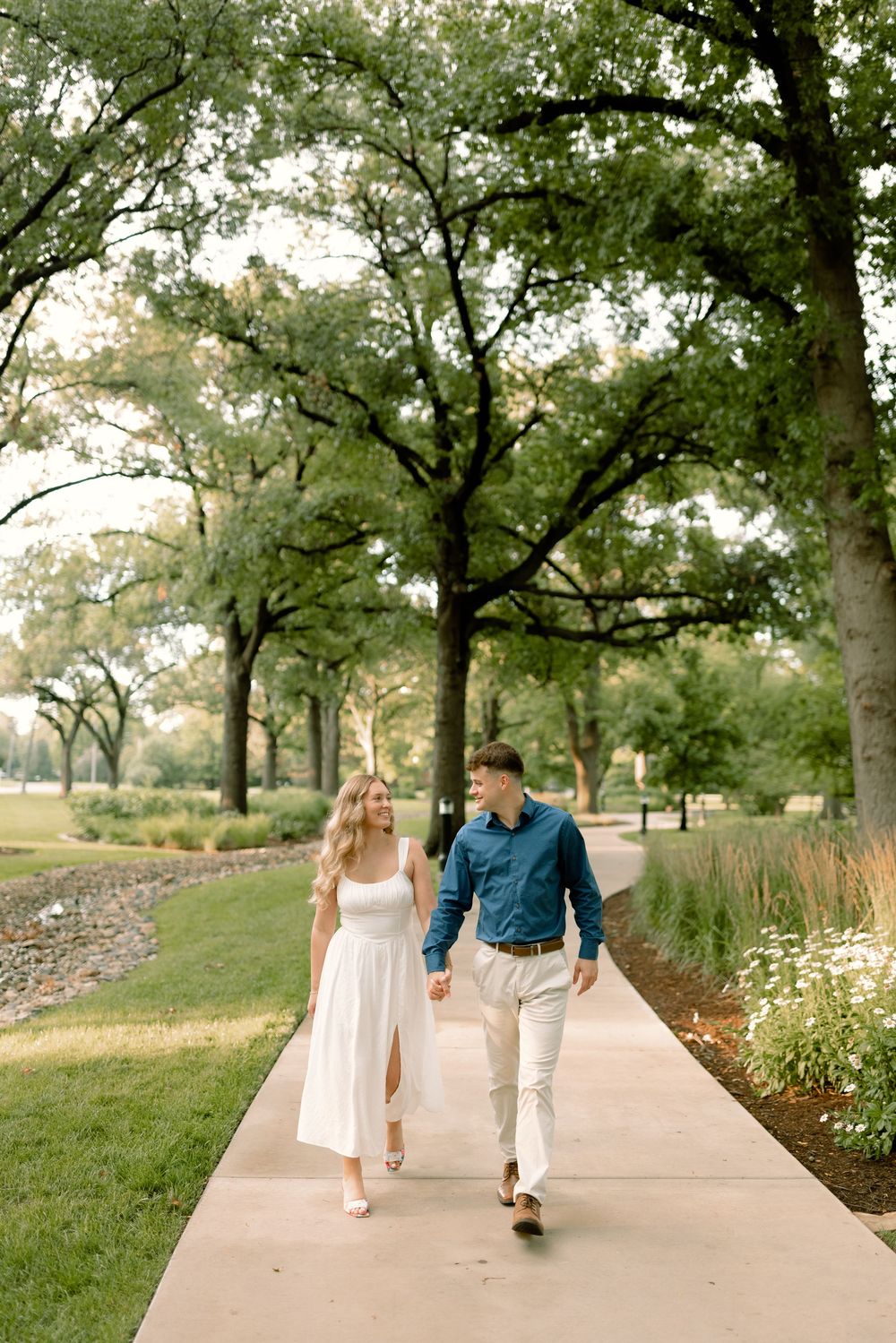 A couple walks hand in hand along a tree-lined path in a park during a summer evening.