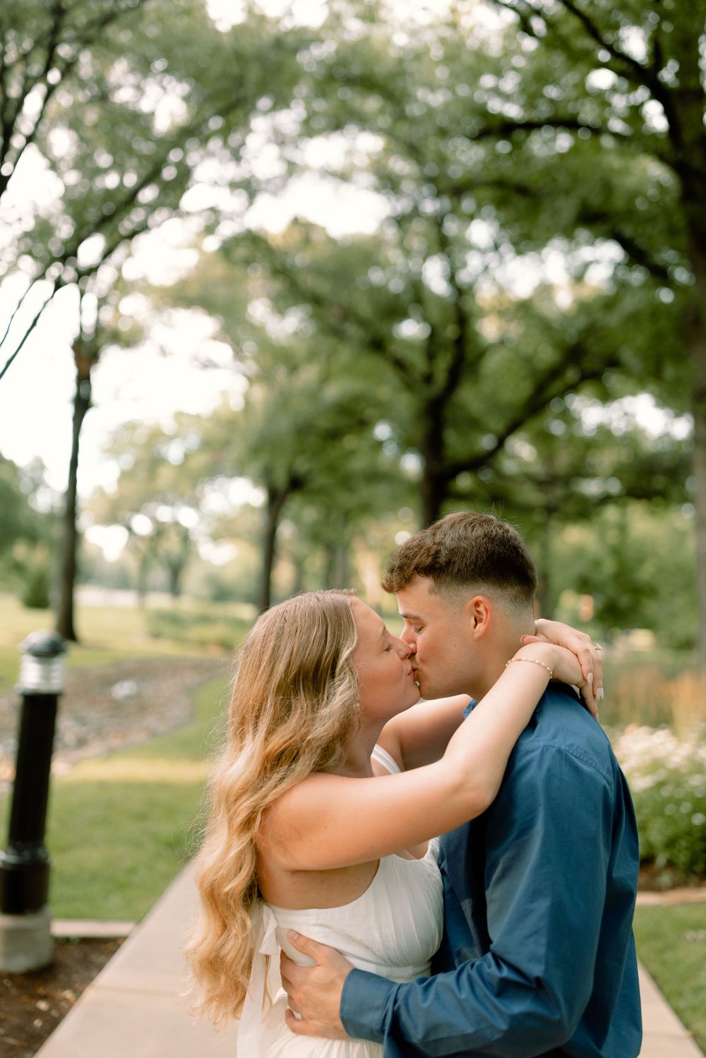 A romantic couple shares tender kisses in a serene outdoor park setting with green trees in the background.