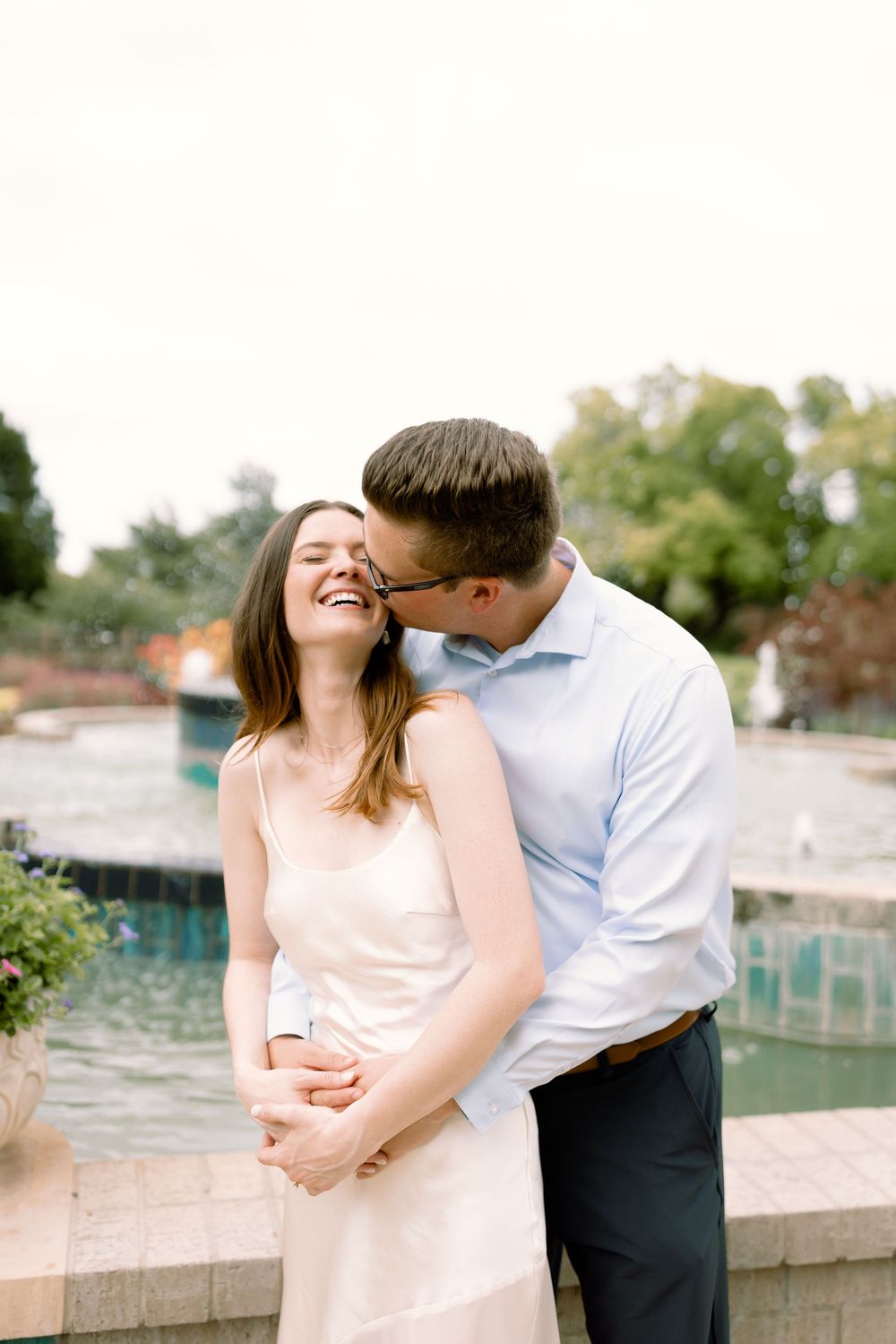 A romantic couple in light pink and white outfits share tender moments along a stone wall during an engagement photoshoot.