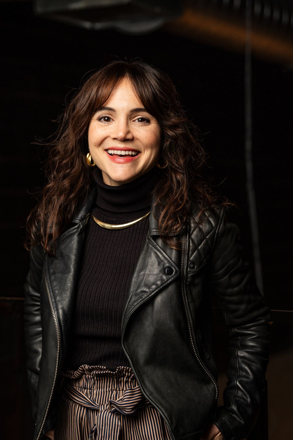 Person in black leather jacket and turtleneck smiling against dark backdrop.