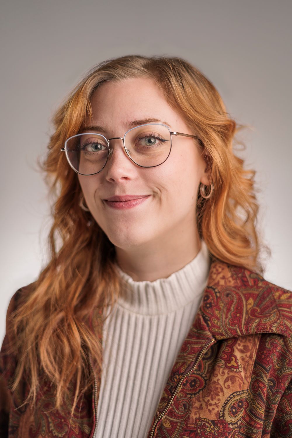 Portrait of someone wearing glasses and a white turtleneck sweater with a paisley print jacket against neutral background.
