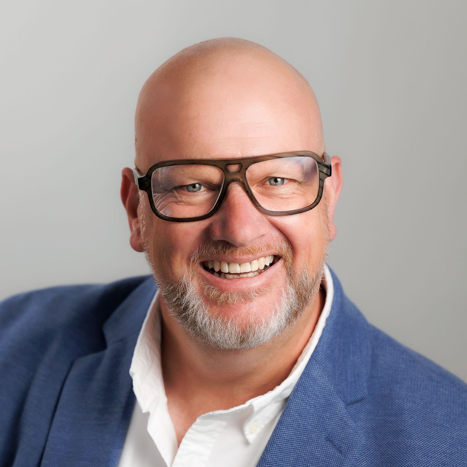 Approachable professional Linkedin headshot of friendly male in blue suit and white shirt smiling warmly while wearing black-framed glasses.