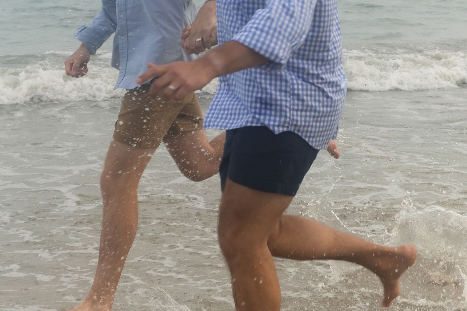 Two husbands holding hands while splashing through shallow ocean waves at Matunuck Beach in South Kingstown RI.