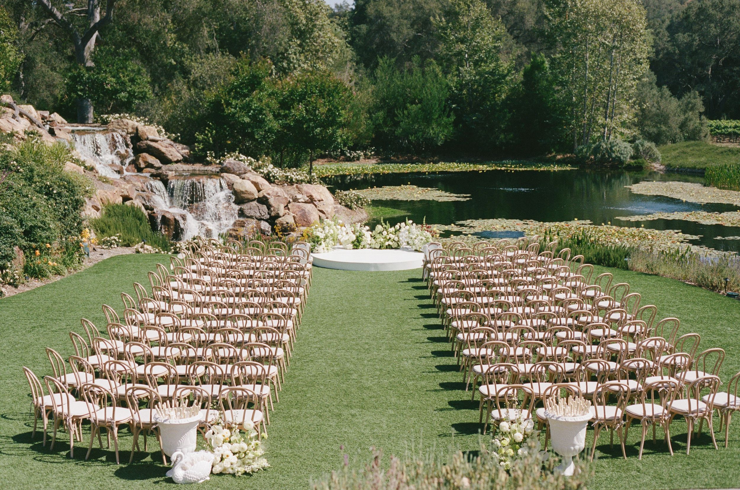 Elegant outdoor wedding ceremony setup with rows of chairs on green lawn, floral altar, pond, and waterfall backdrop.