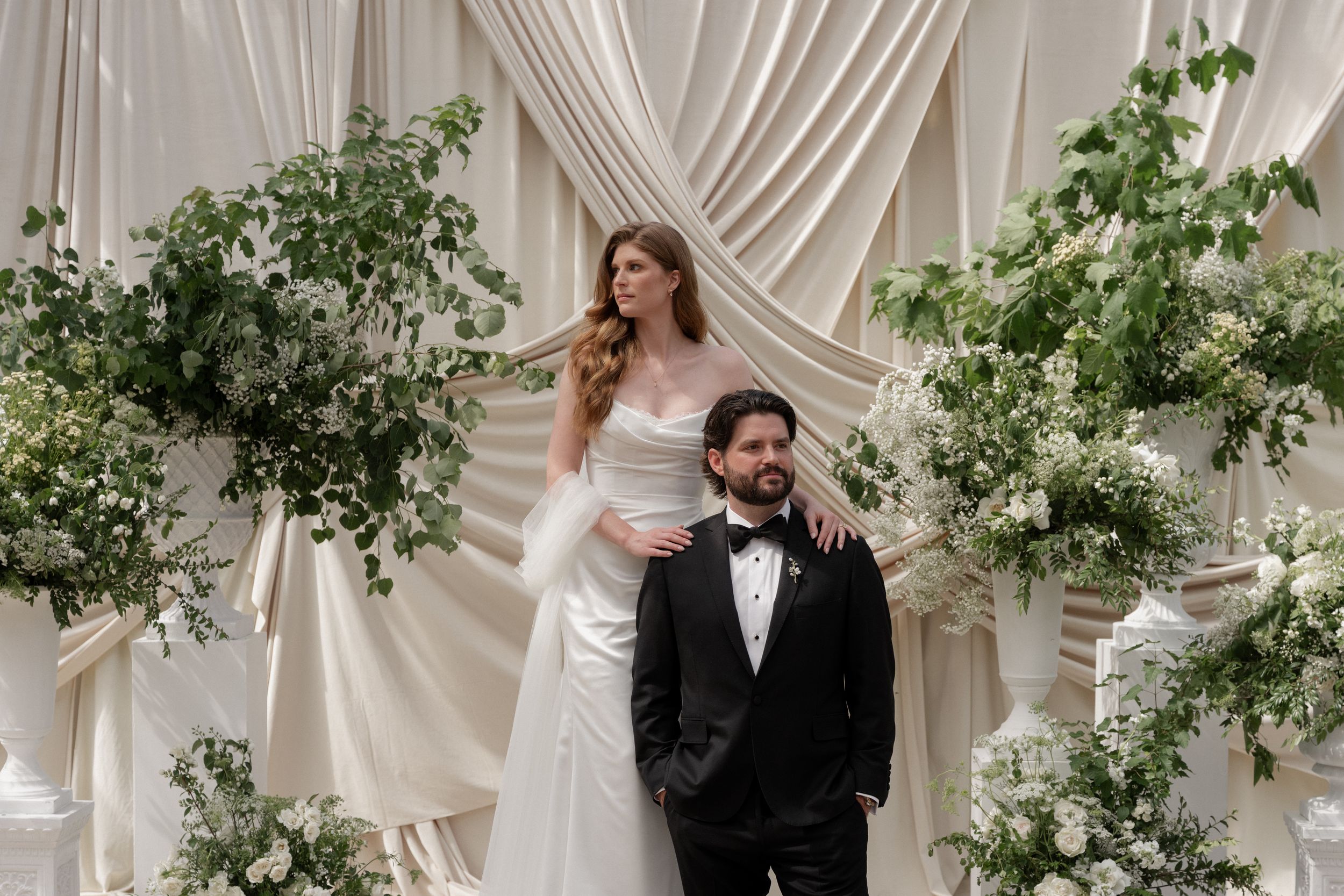 Elegant bride and groom pose before lush floral arch with white draping and greenery at luxury wedding ceremony.