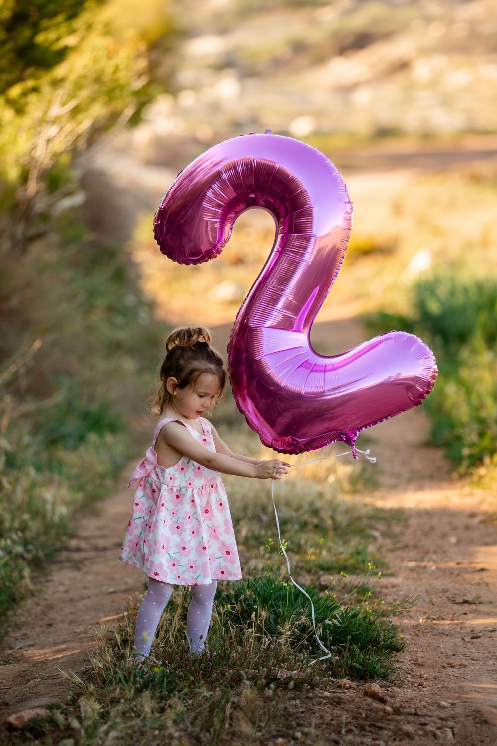 Young child in dress poses with pink number balloon outdoors.