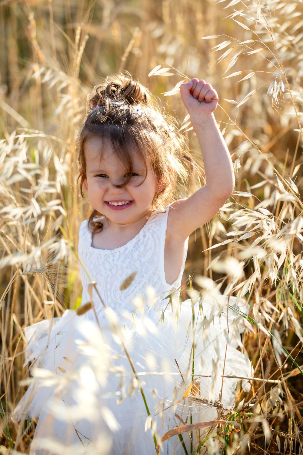 Child in white dress plays joyfully among tall grass in sunlight.