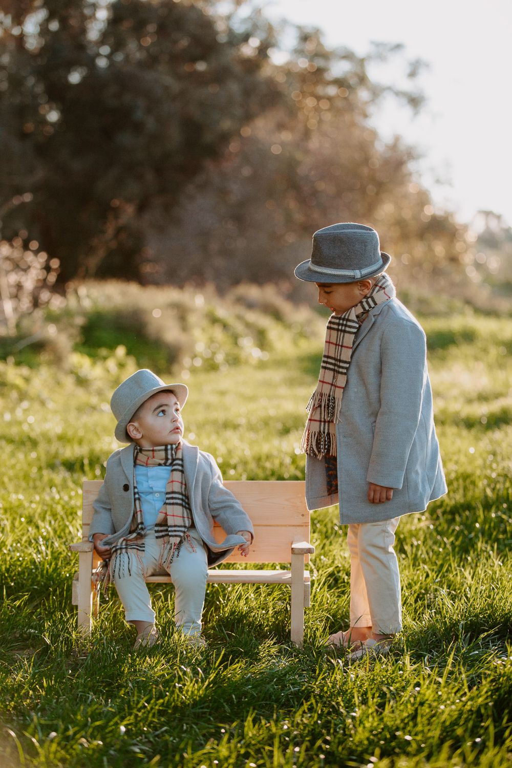 Two small figures in matching gray coats, scarves and hats stand together by a bench in a sunlit meadow at golden hour.