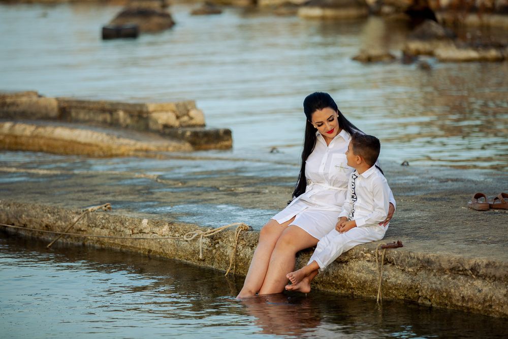 Two people in white outfits sit together on rocky coastal ledge during sunset engagement photoshoot at the beach.