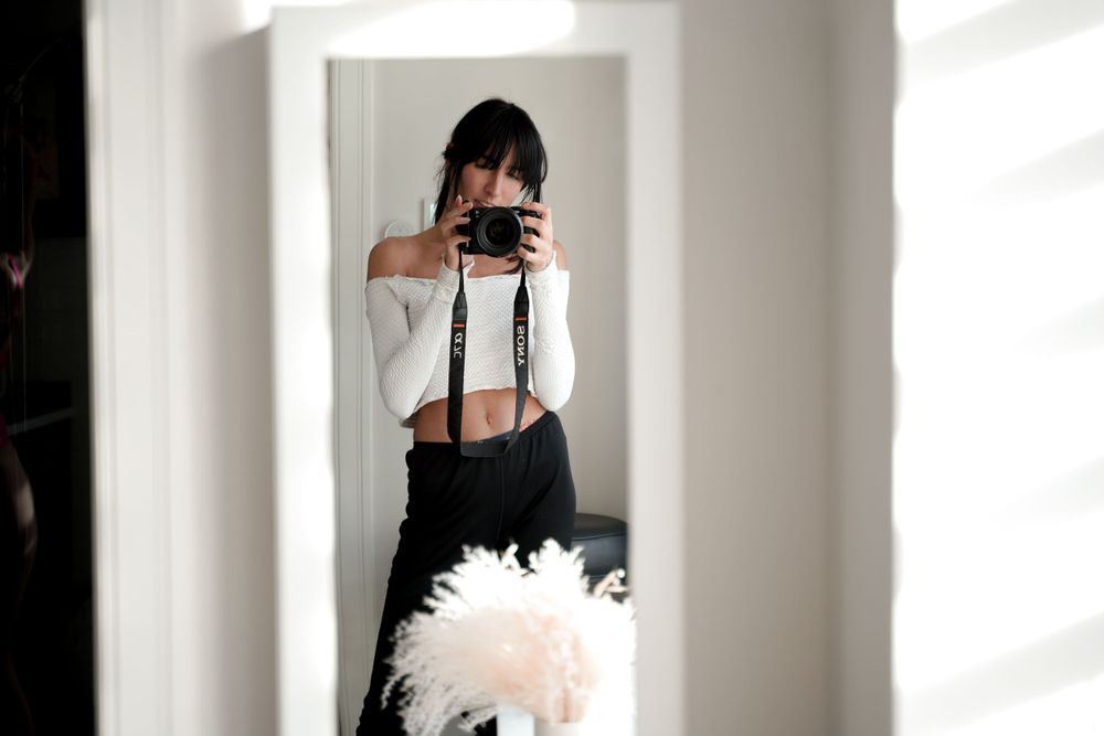 Mirror selfie showing person with camera in white top and dark pants.