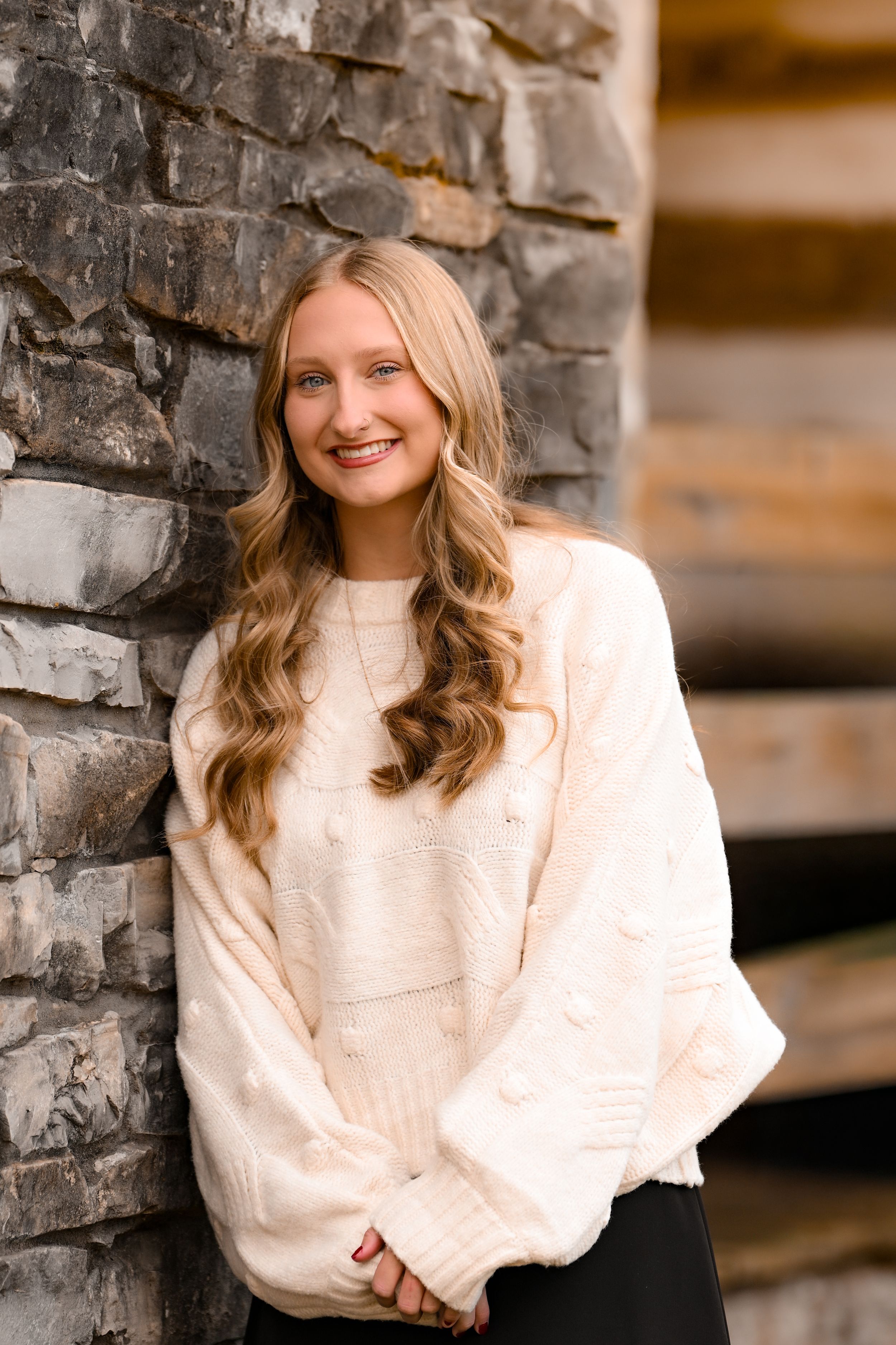 Young woman in cream sweater smiling while leaning against stone wall.