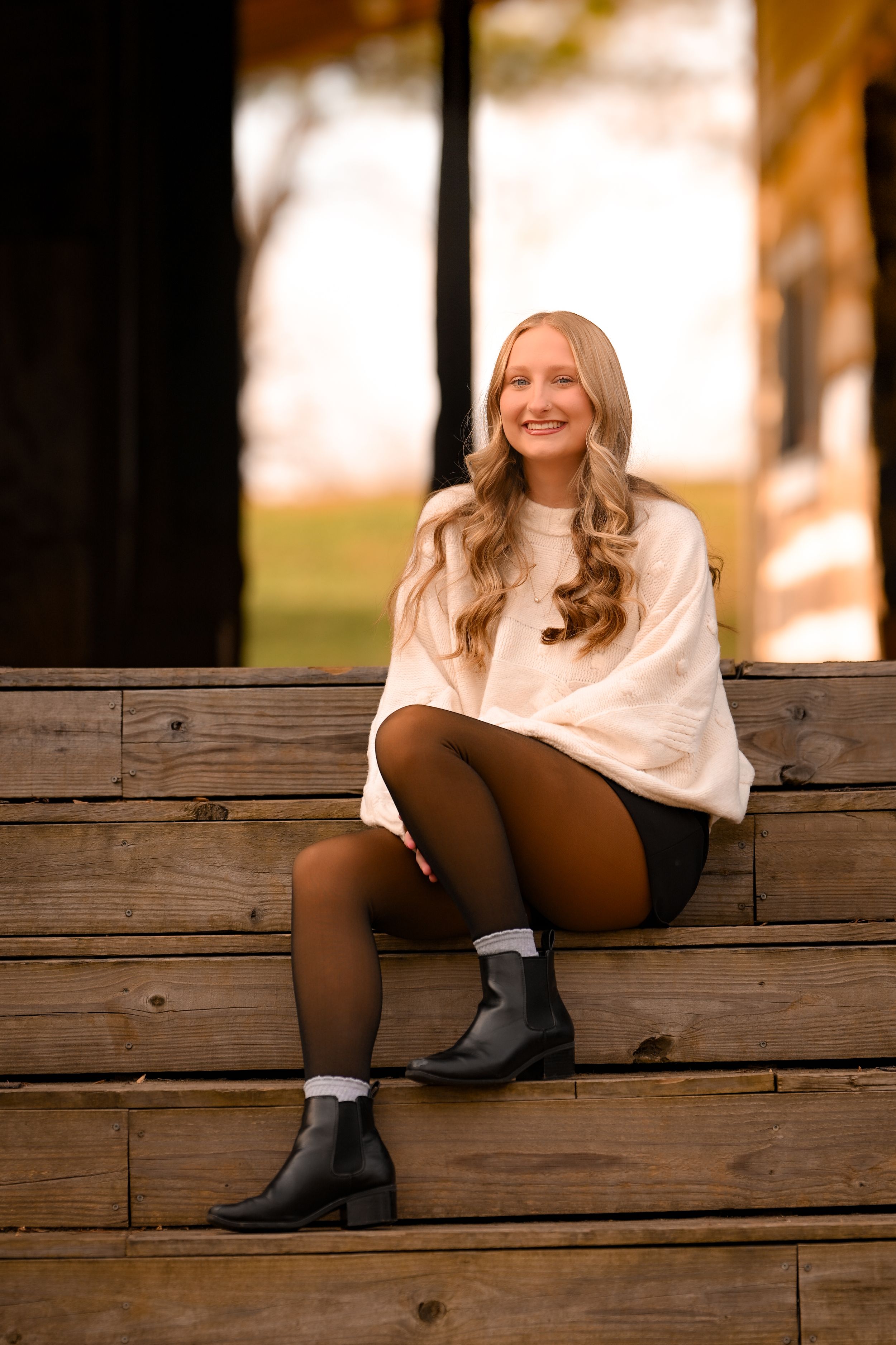 Woman in white sweater sitting on wooden steps wearing black boots and tights.